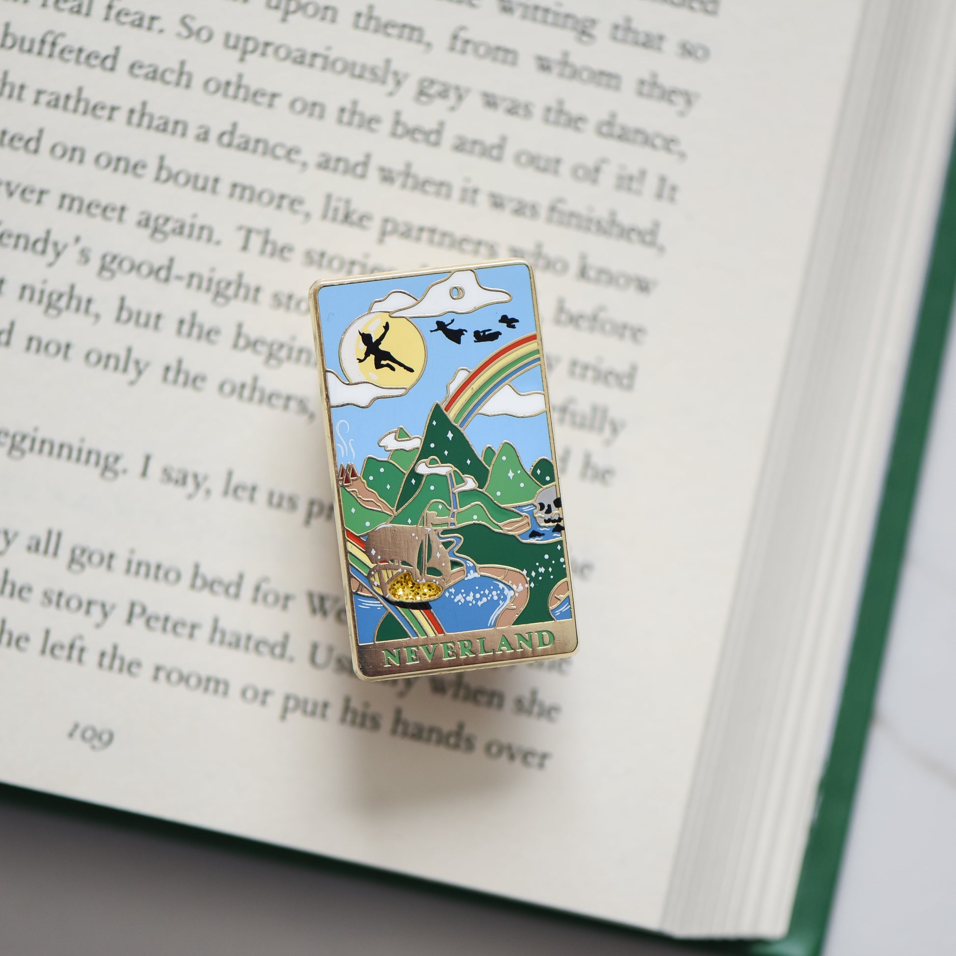 Gold Neverland landscape enamel pin with rainbow, moon, peter pan silhouette, and fairy ship details on an open book