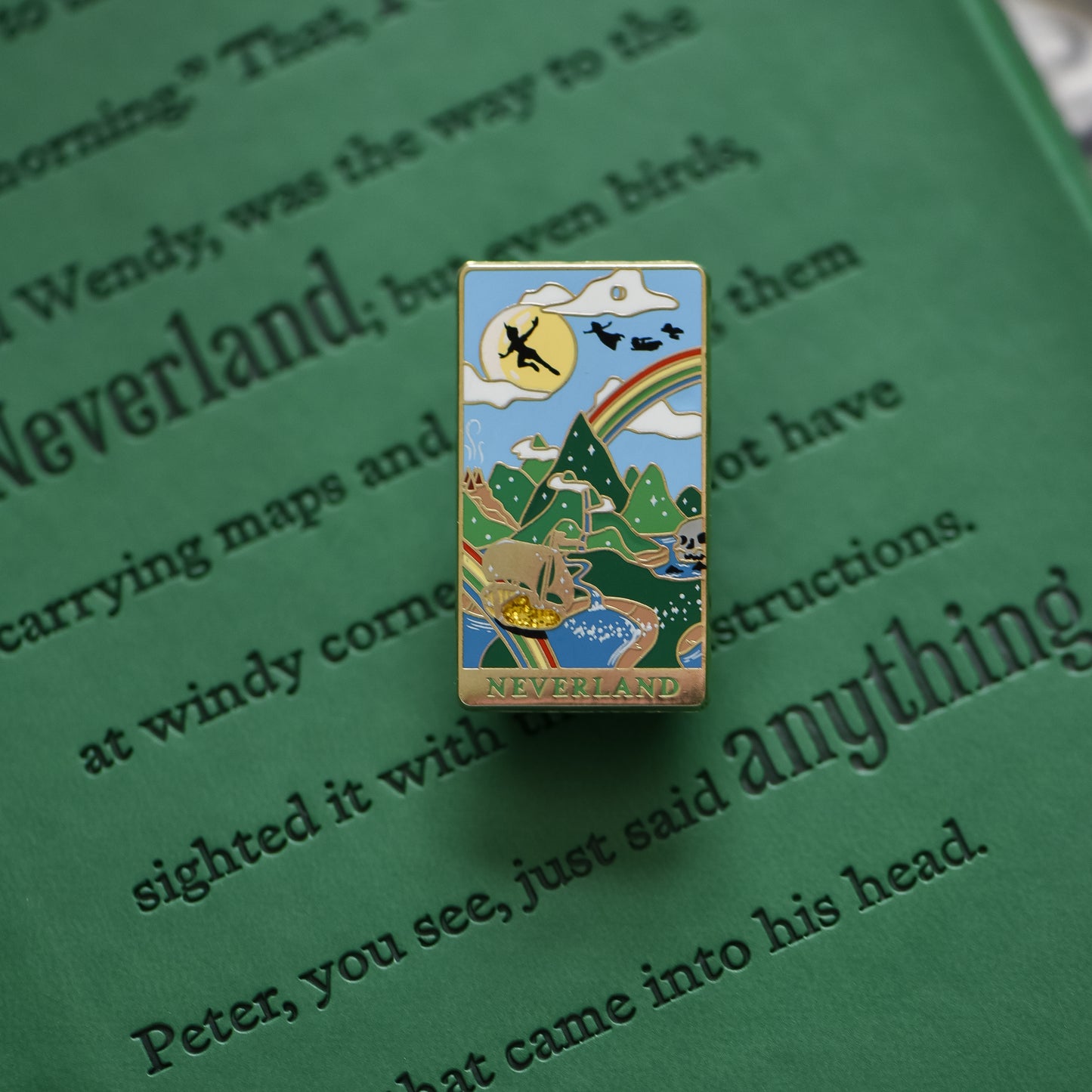 Gold Neverland landscape enamel pin with rainbow, moon, peter pan silhouette, and fairy ship details on green book