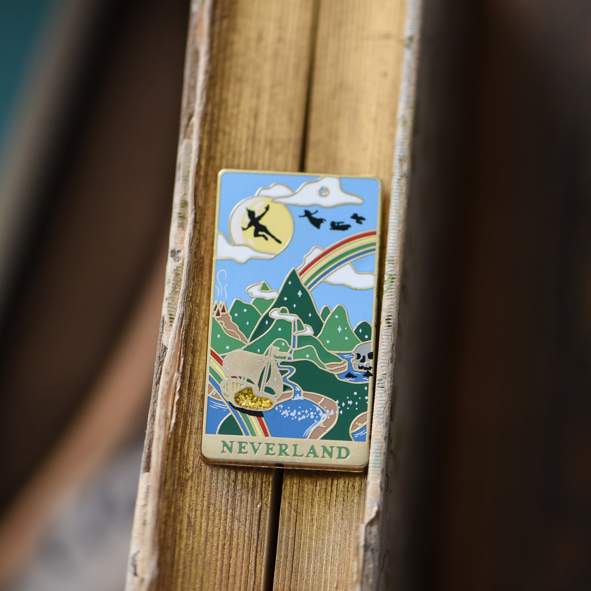Gold Neverland landscape enamel pin with rainbow, moon, peter pan silhouette, and fairy ship details on an open book