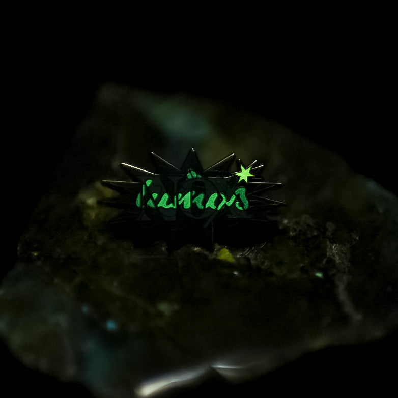 Black nickel enamel pin with Nox in green letters with glow in the dark lumos letters on a potion spell book