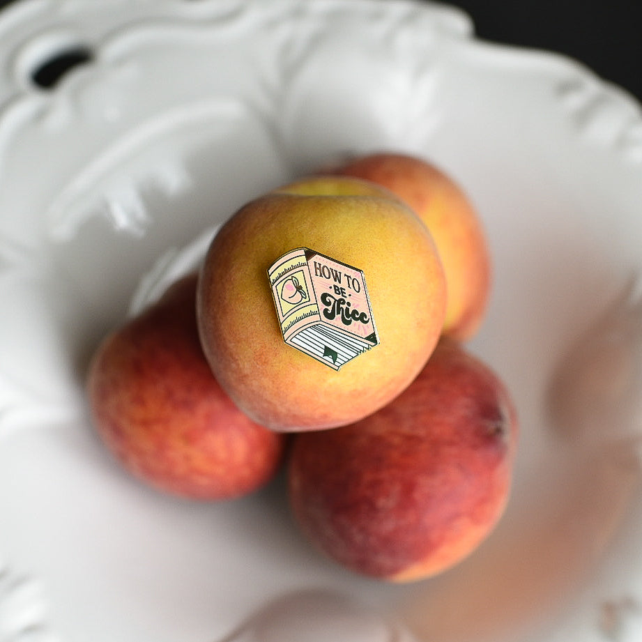 Peach book enamel pin with the title How to be Thicc on a stack of peaches