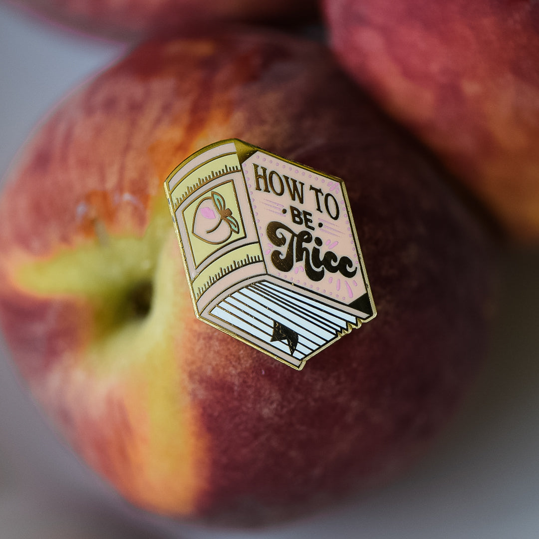 Peach book enamel pin with the title How to be Thicc on a stack of peaches