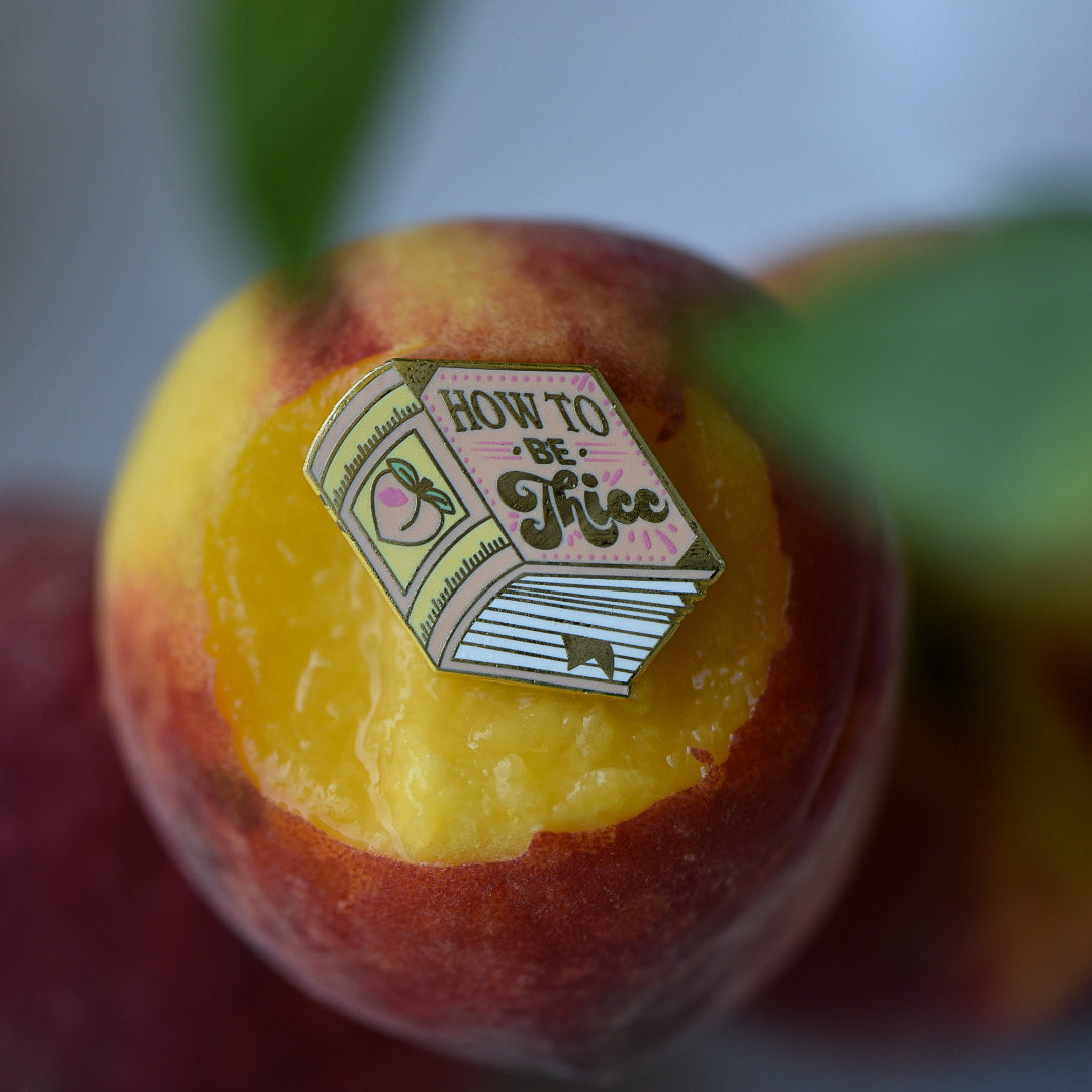 Peach book enamel pin with the title How to be Thicc on a stack of peaches