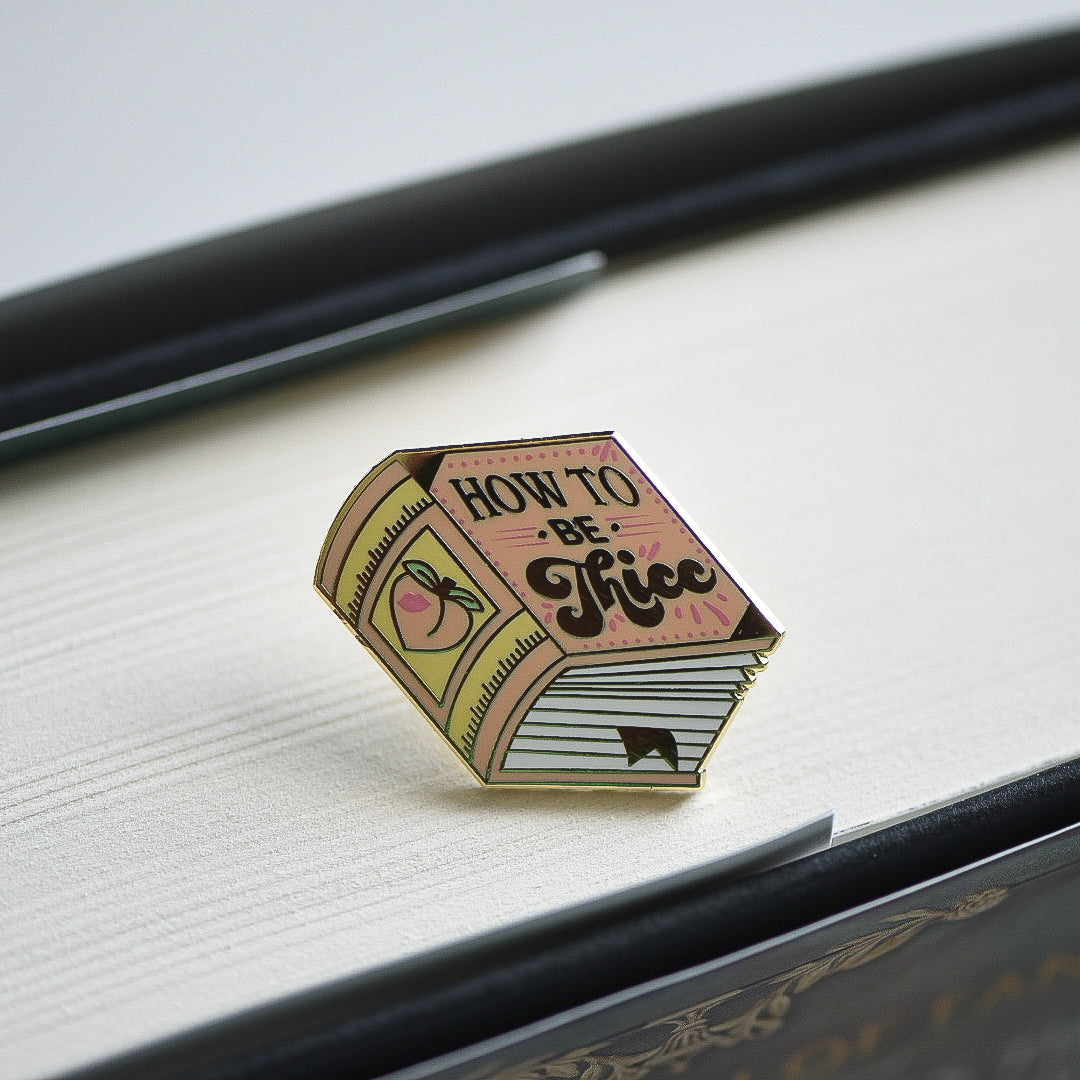 Peach book enamel pin with the title How to be Thicc on an open book