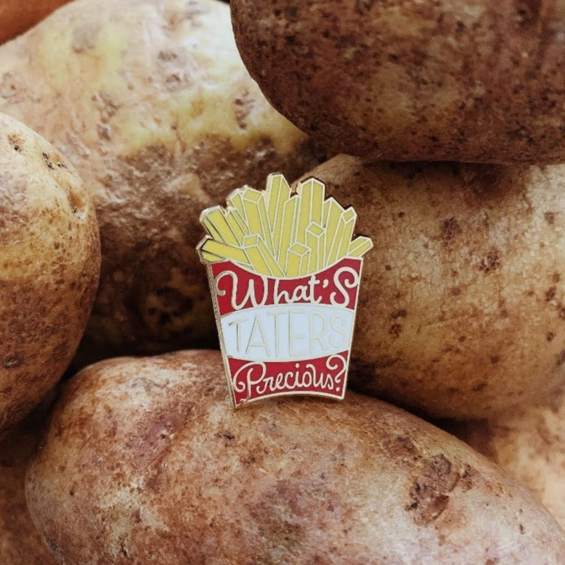 Red box of fries gold enamel pin that says What's Taters Precious on a pile of potatoes