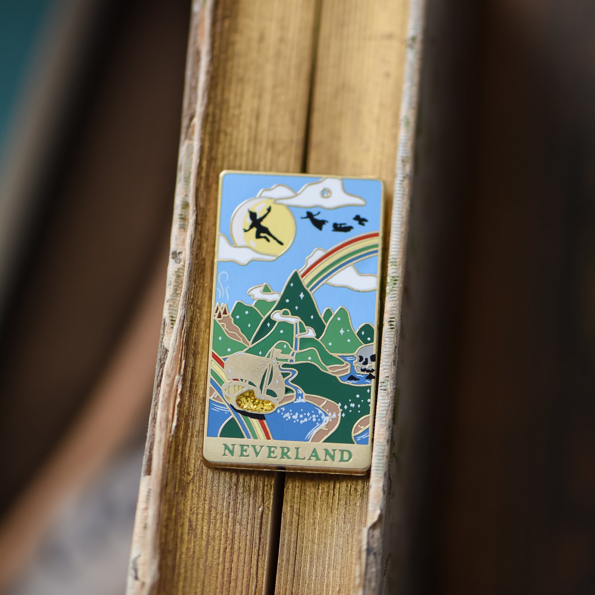 Gold Neverland landscape enamel pin with rainbow, moon, peter pan silhouette, and fairy ship details on an open book