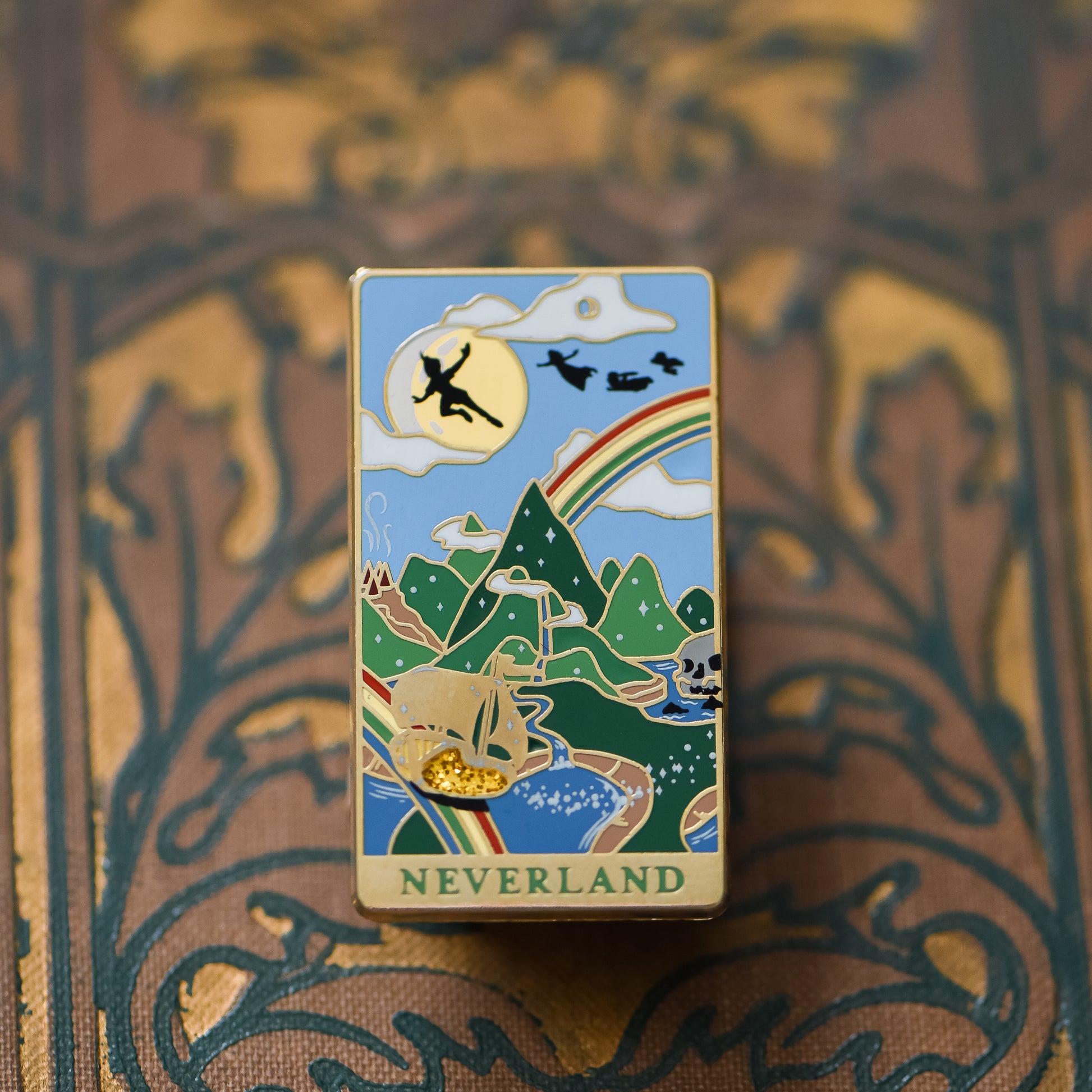 Gold Neverland landscape enamel pin with rainbow, moon, peter pan silhouette, and fairy ship details on an open book