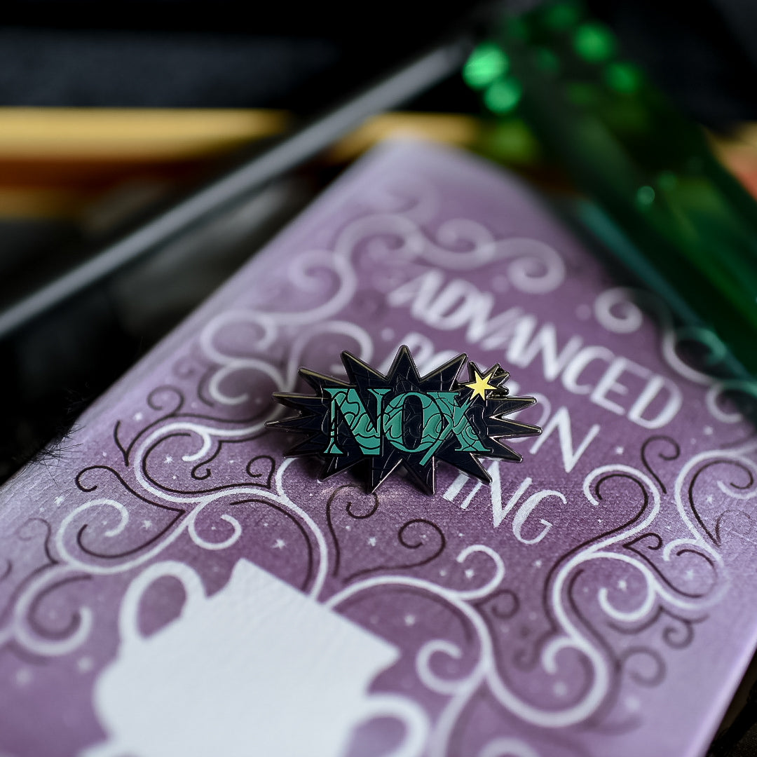 Black nickel enamel pin with Nox in green letters with glow in the dark lumos letters on a potion spell book
