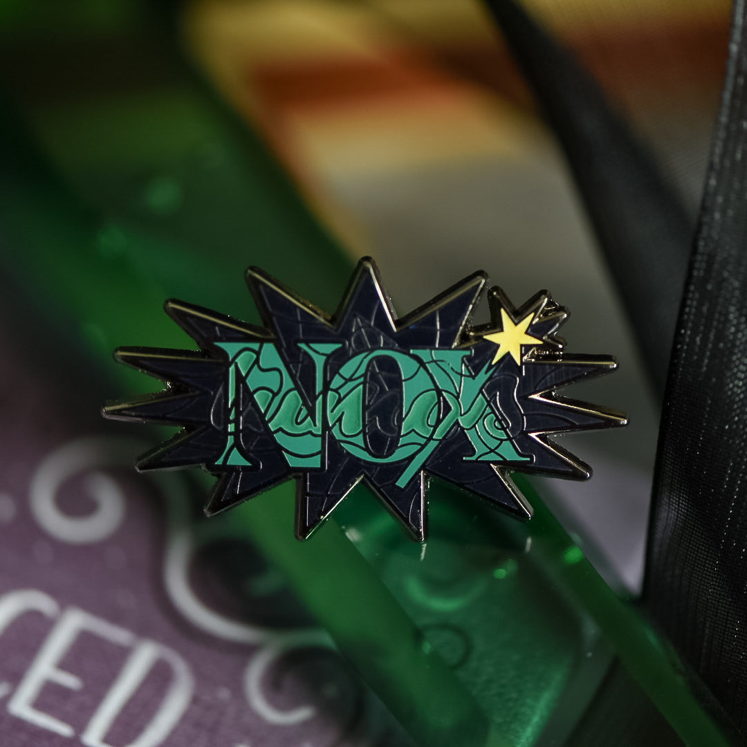 Black nickel enamel pin with Nox in green letters with glow in the dark lumos letters on a potion spell book