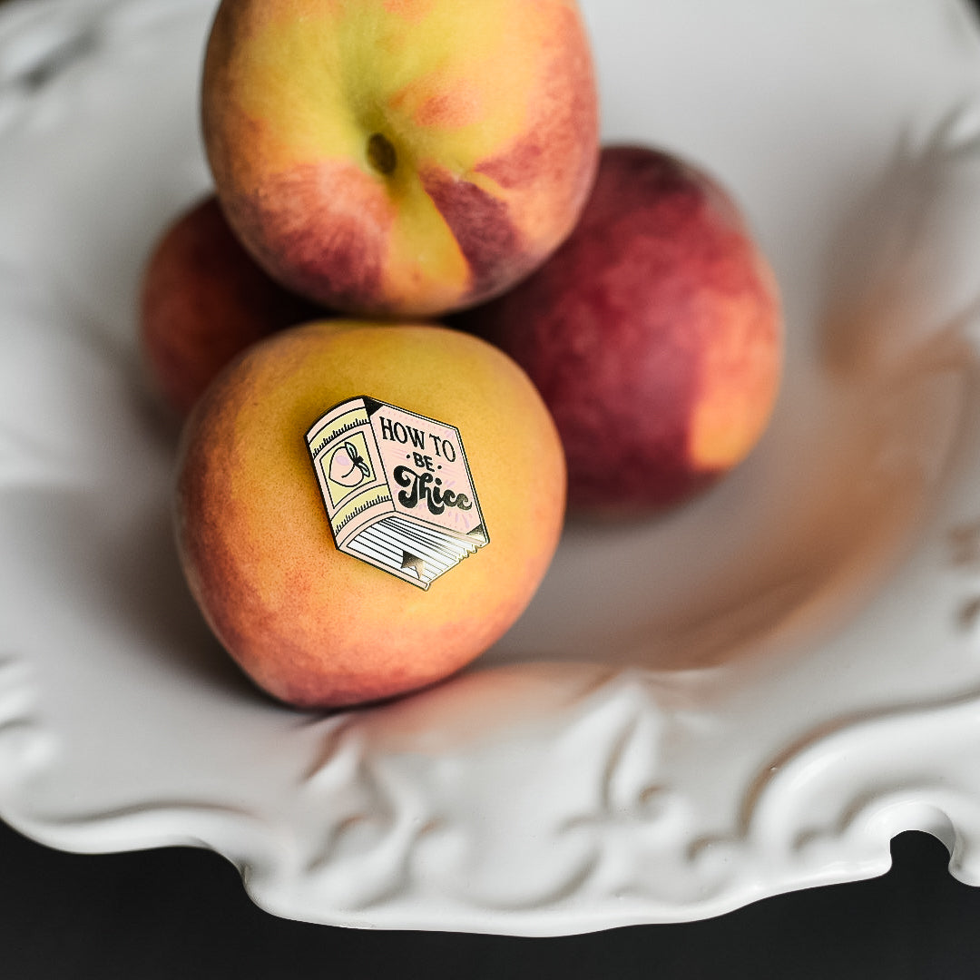 Peach book enamel pin with the title How to be Thicc on a stack of peaches