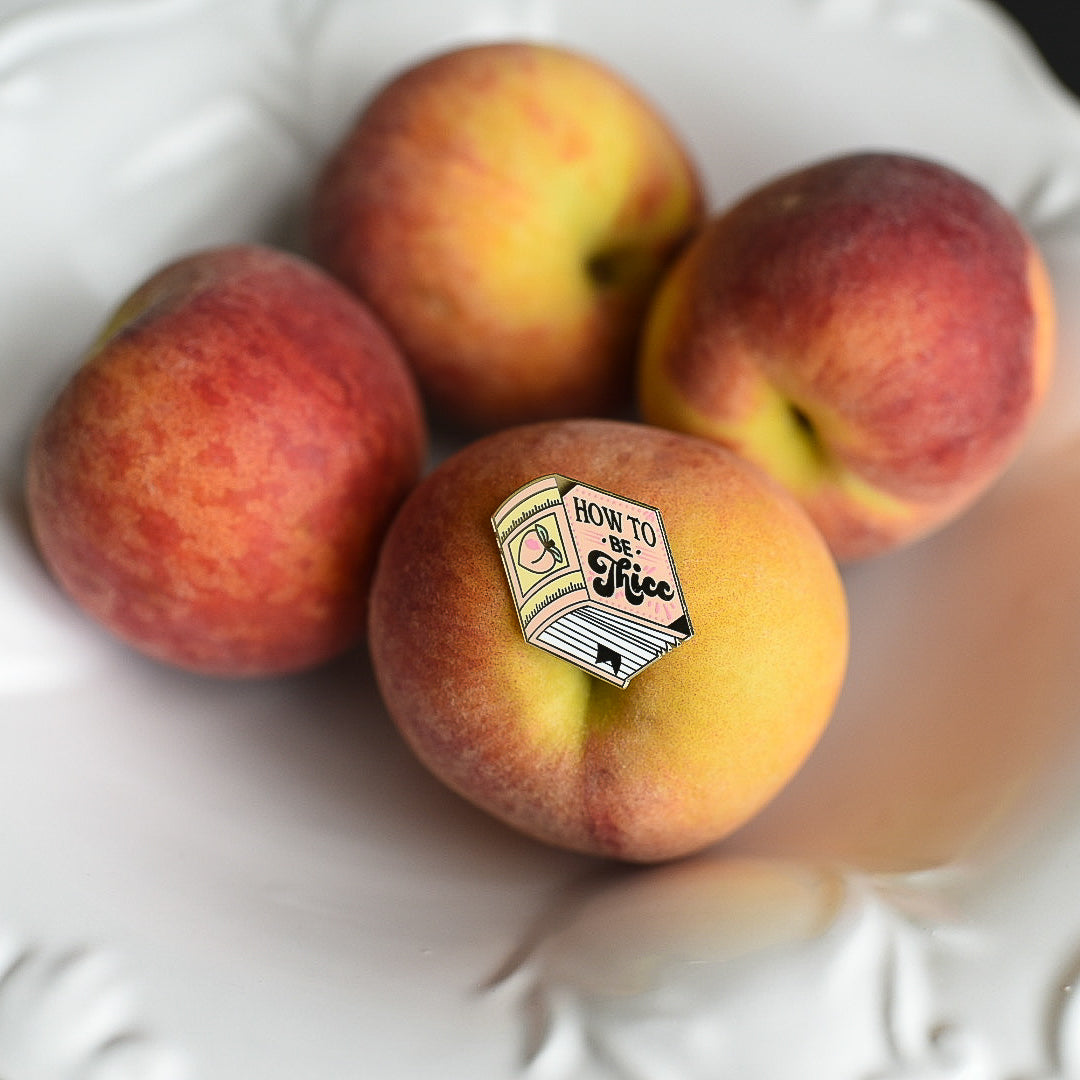 Peach book enamel pin with the title How to be Thicc on a stack of peaches