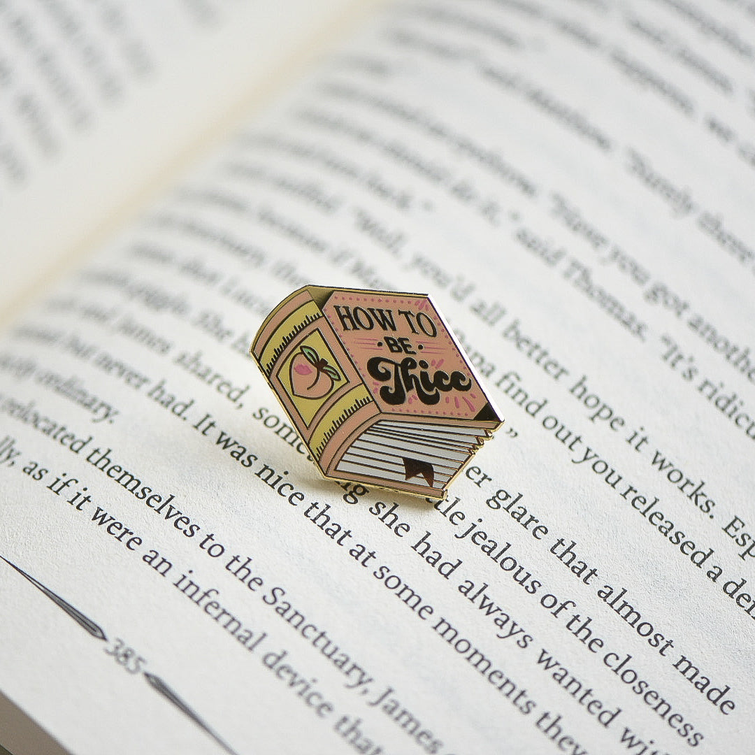 Peach book enamel pin with the title How to be Thicc on an open book