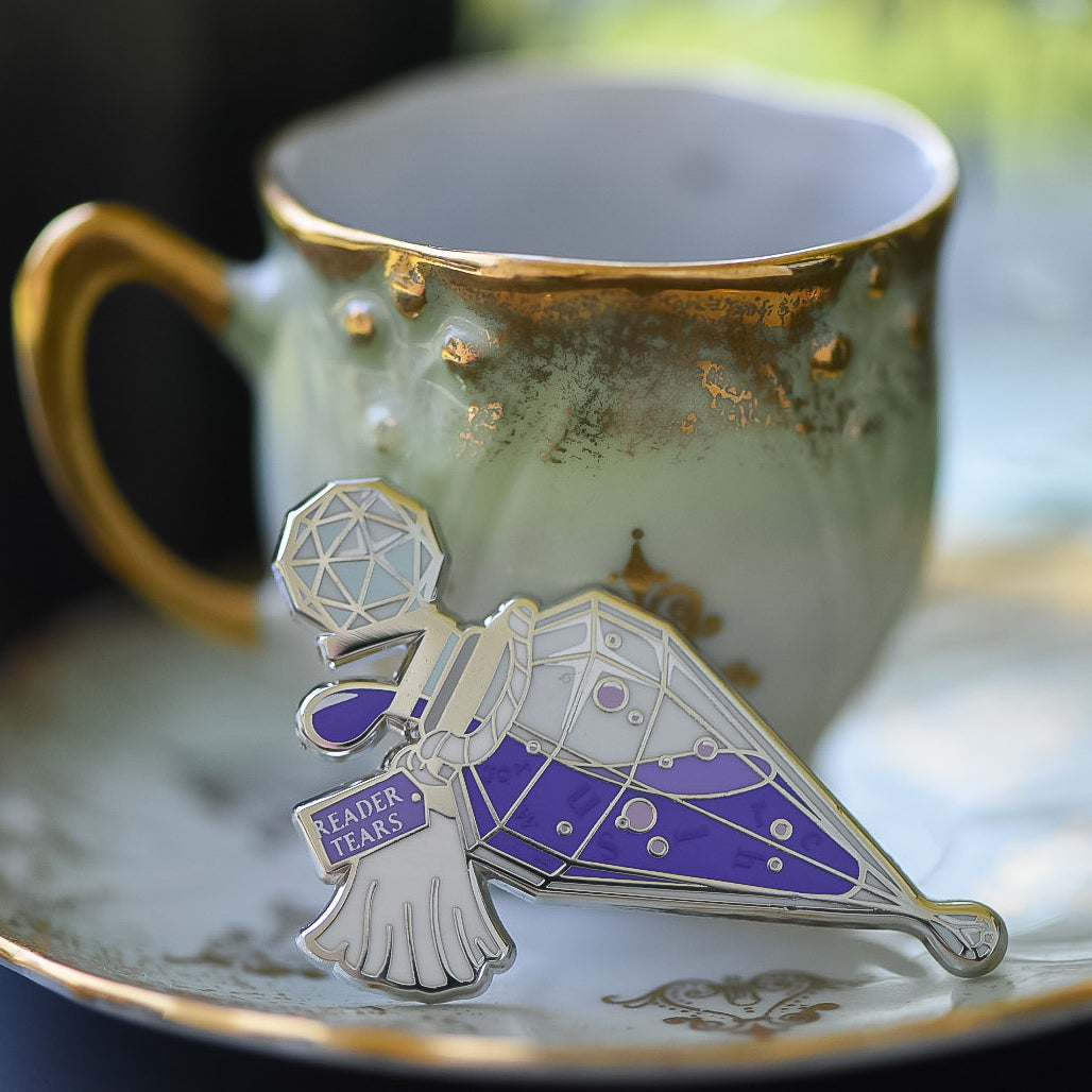 Purple and silver potion bottle enamel pin with a tag that says Reader Tears sitting on a teacup