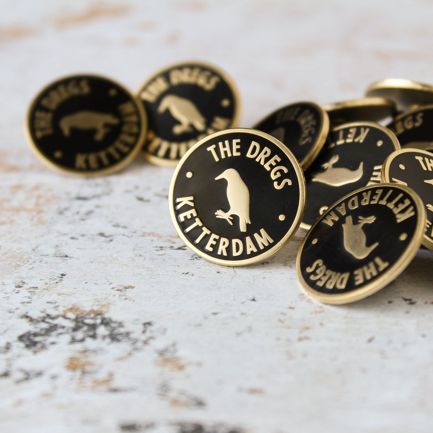 Pile of black and matte gold dregs membership style enamel pin on a white background