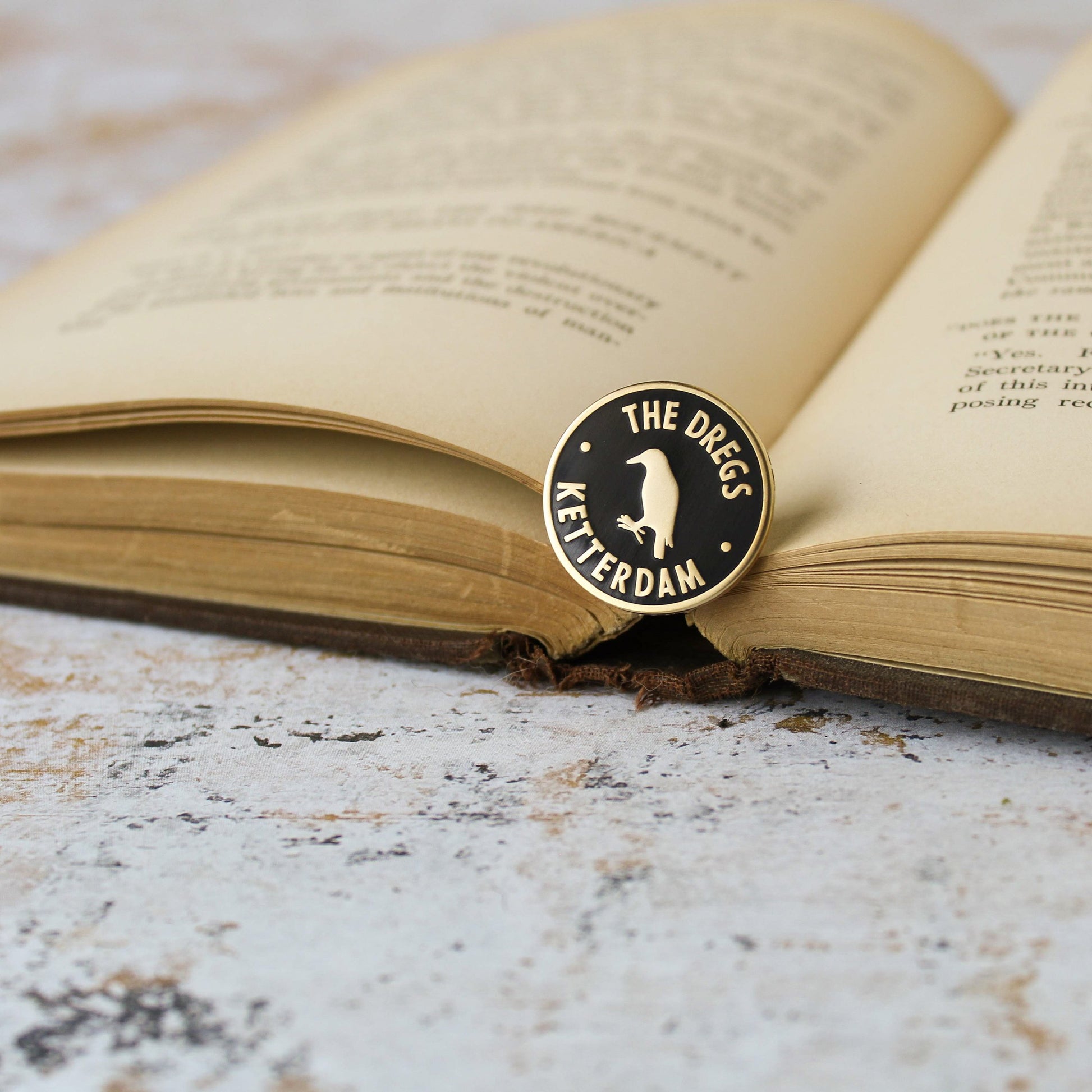 Black and matte gold crow inside a circle dregs membership style enamel pin on an open book