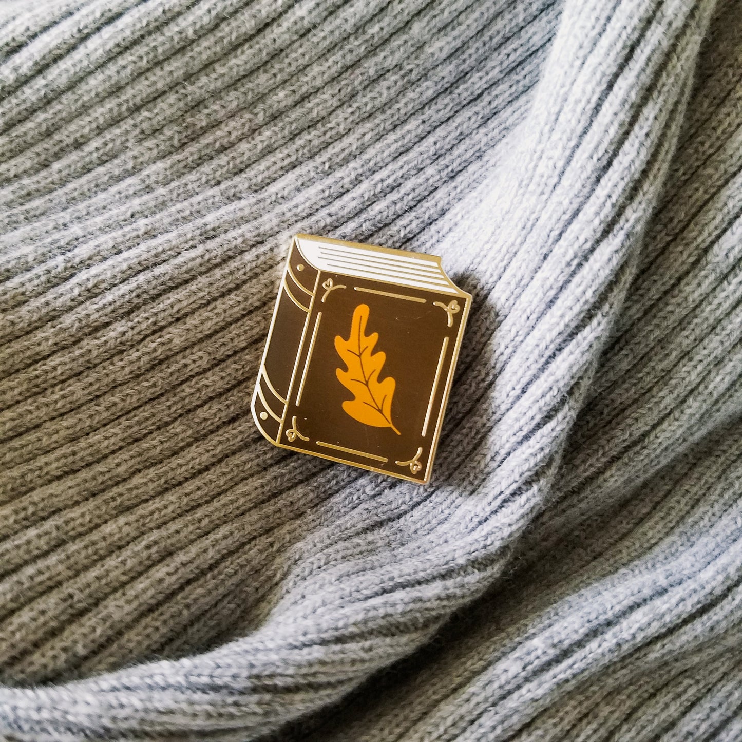 Chocolate brown book with small orange leaf enamel pin resting on a book