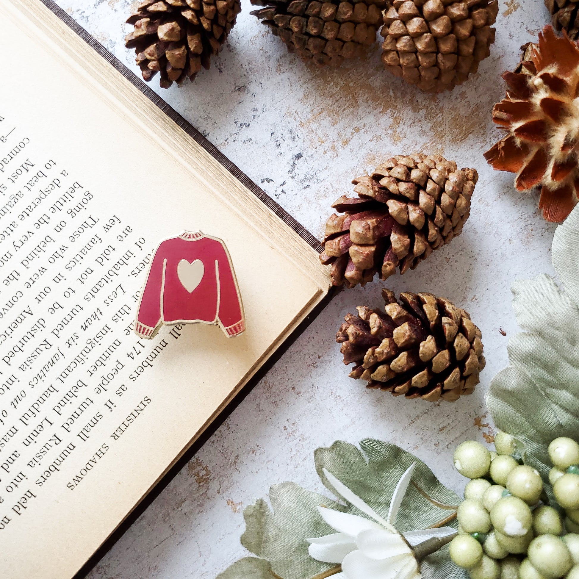 Maroon red hand stamped heart sweater enamel pin with a book and pine cones