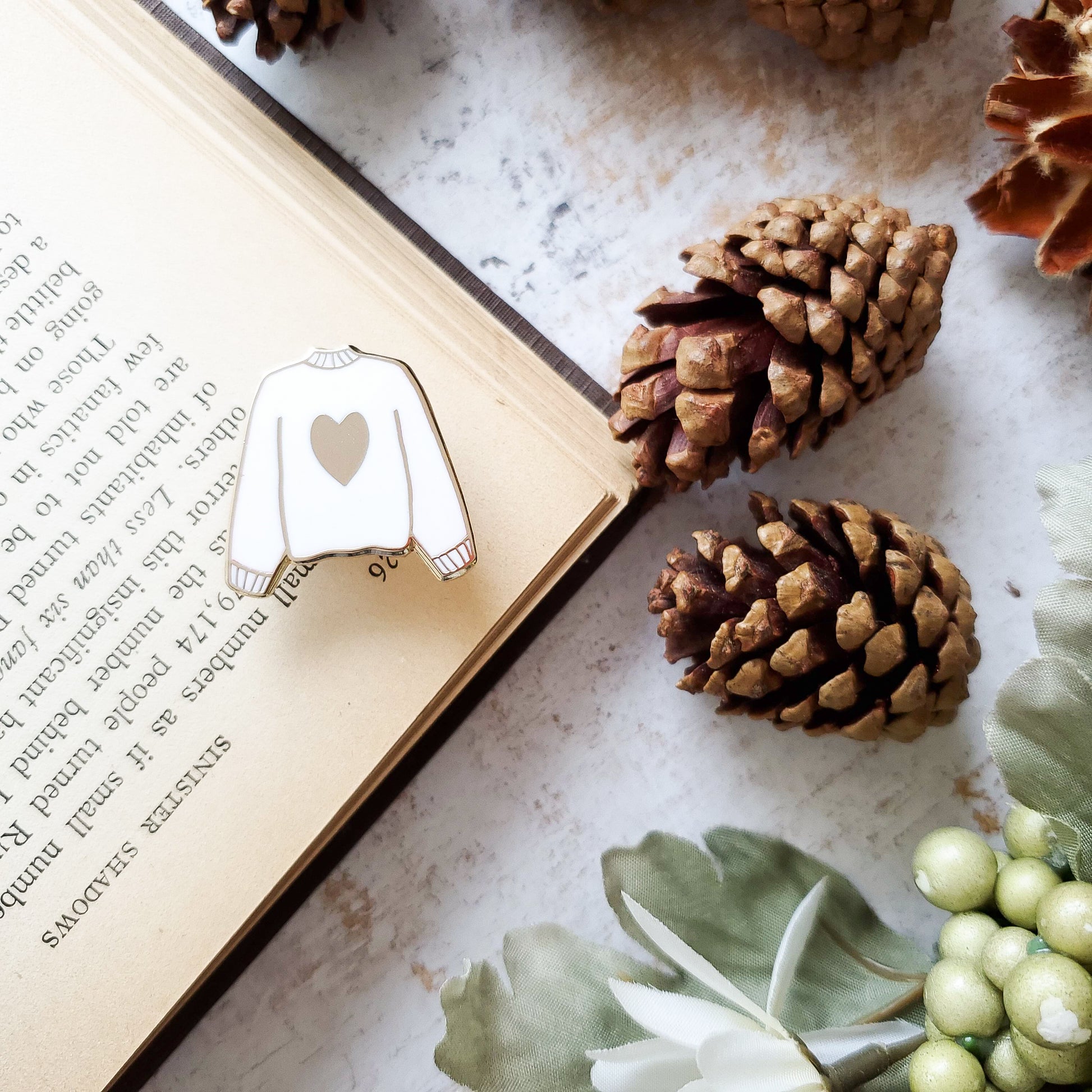 White hand stamped heart sweater enamel pin with a book and pine cones