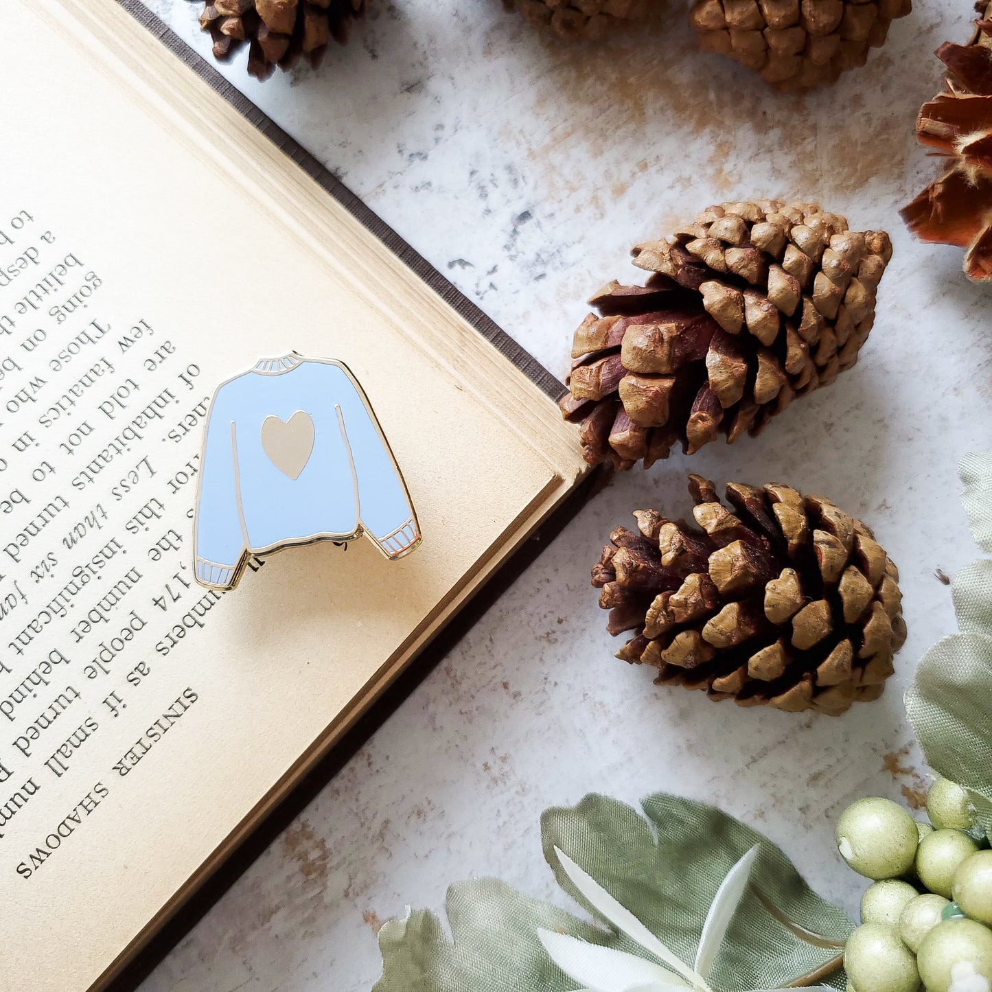 Blue hand stamped heart sweater enamel pin with a book and pine cones