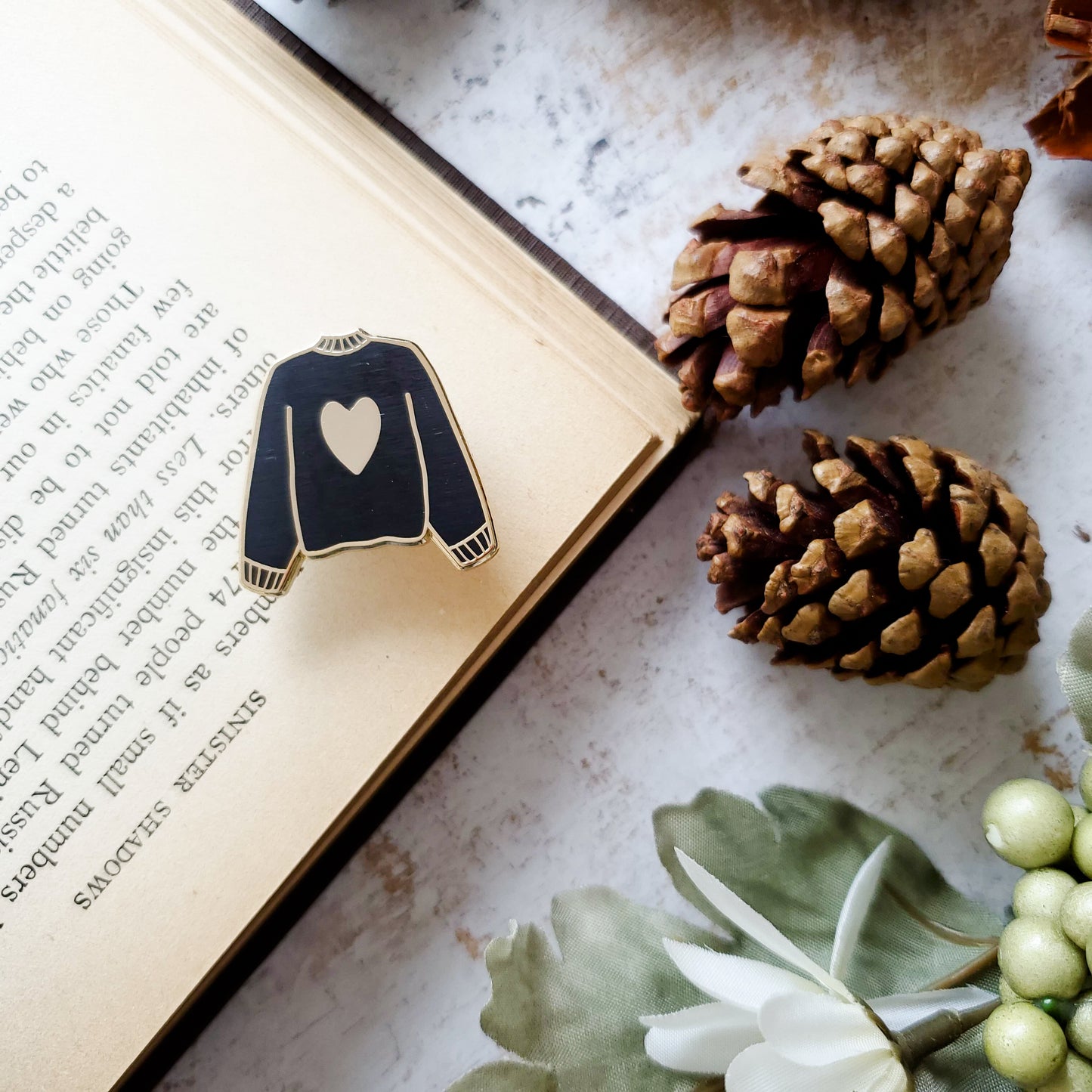 Black hand stamped heart sweater enamel pin with a book and pine cones