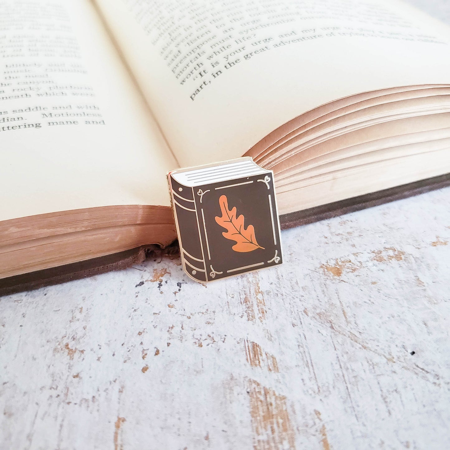 Chocolate brown book with small orange leaf enamel pin resting on a book